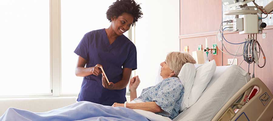A black nurse with a smile on her face shares a tablet with a white elderly patient lying in a hospital bed.